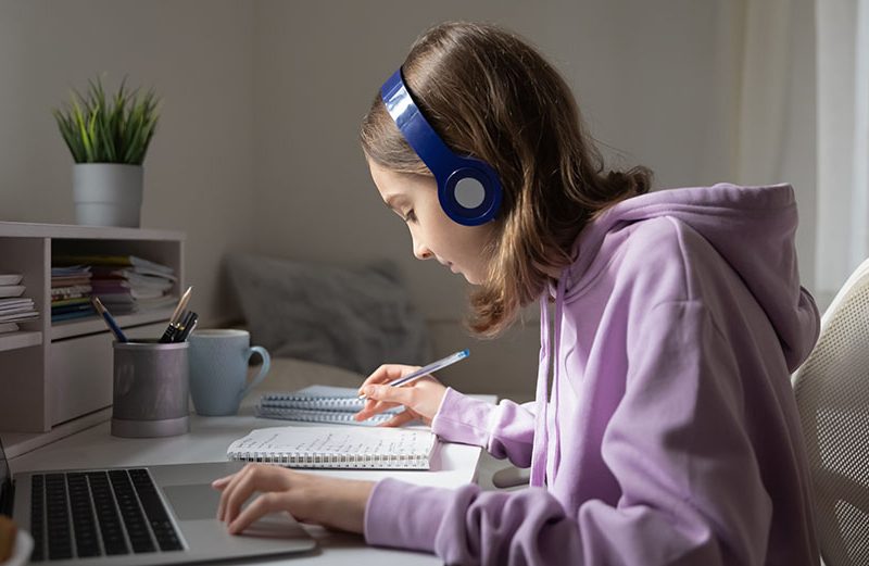 Teen girl school pupil wearing headphones studying online from home making notes. Teenage student distance learning on laptop doing homework, watching listening video lesson. Remote education concept